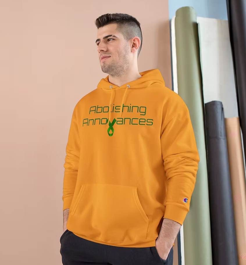 A person wears an orange hoodie with green text on the front reading “Abolishing Annoyances.” The word “Abolishing” appears above “Annoyances,” with the letter “i” stylized as a green ribbon. The hoodie features a front pocket and drawstrings, and the person stands against a pink wall with a partially visible curtain. The design blends bold messaging with symbolic advocacy, suggesting a stance against everyday stressors or mental health stigma.