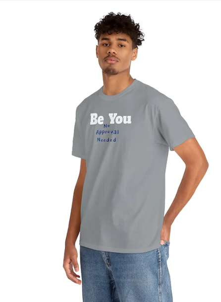 A person wears a gray t-shirt paired with blue jeans. The shirt features a bold, motivational message: “Be You / No / Approval / Needed,” arranged in stacked lines across the front. The typography emphasizes self-acceptance and confidence, encouraging individuality without external validation. The overall look conveys strength through simplicity and personal empowerment.