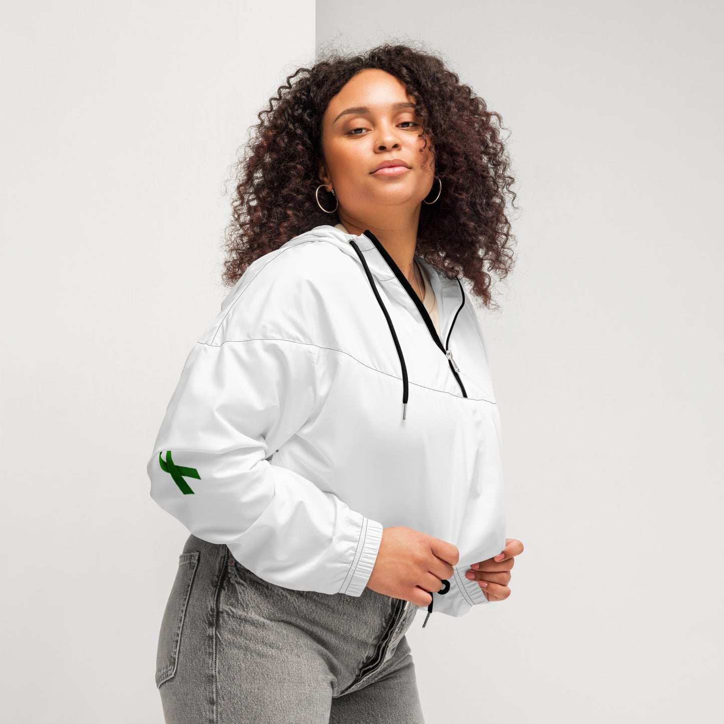 A person poses confidently in a white zip-up pullover with a black zipper and drawstring at the waist, paired with high-waisted gray jeans. The pullover features a small green design on the left sleeve. The individual has curly hair and holds the collar with both hands, standing against a plain background. The outfit blends casual comfort with modern style, subtly hinting at advocacy through the green sleeve detail.