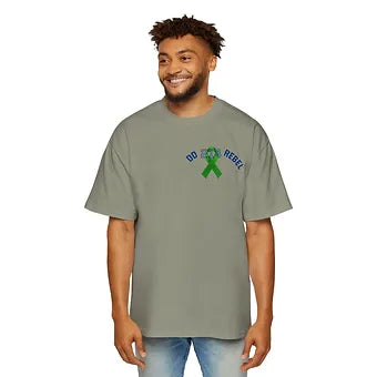 A person wears a grayish-green t-shirt with a small chest design featuring a green awareness ribbon centered over a skull graphic. Above and below the ribbon, the words “OD REBEL” appear in bold blue capital letters. The shirt’s design suggests advocacy around overdose awareness or substance abuse, blending symbolism with a message of resistance and visibility.