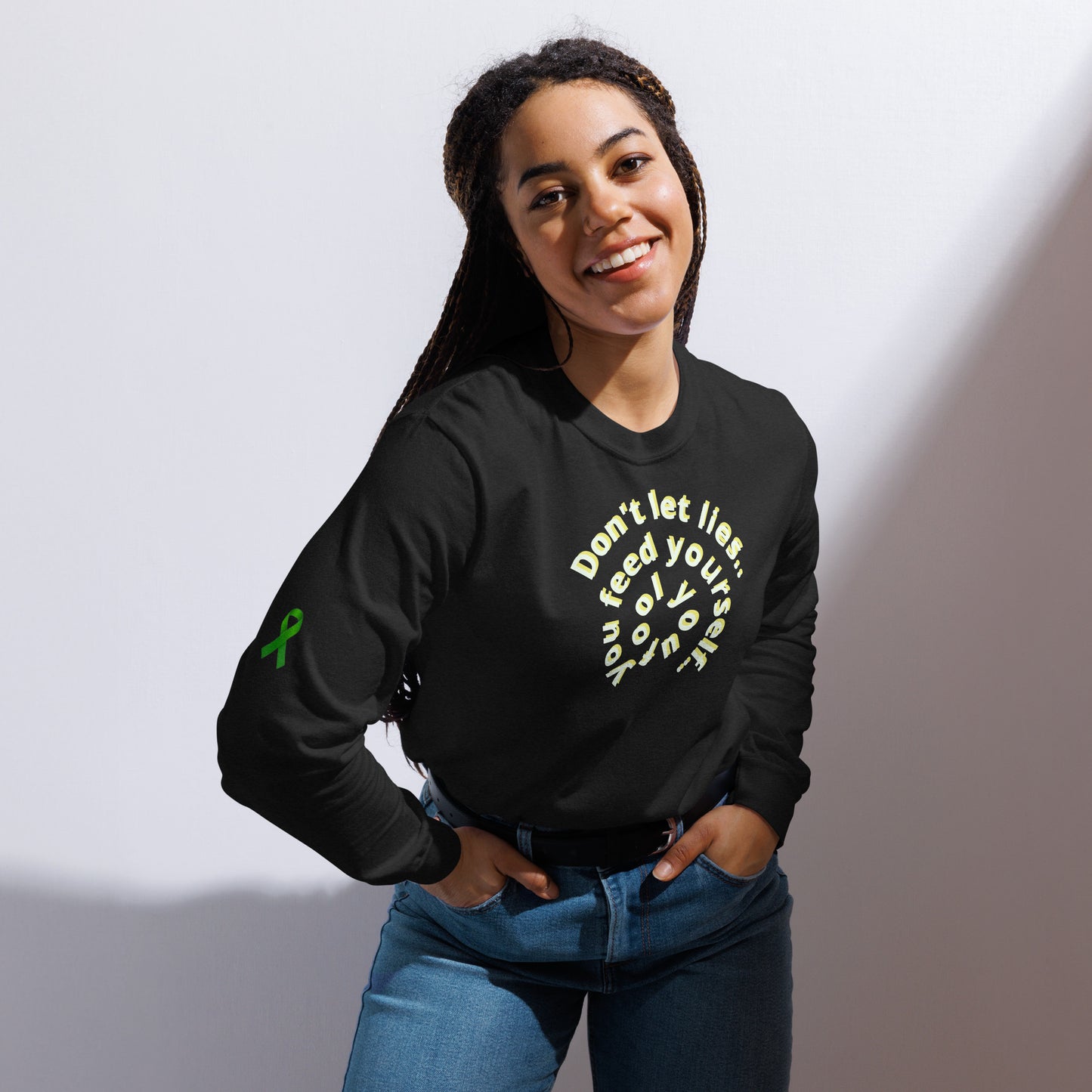 A person stands against a plain background wearing a black long-sleeve shirt with a circular front design that reads “Don’t let illness feed your self-doubt.” A green awareness ribbon is printed on the left sleeve. The shirt’s message promotes mental health encouragement and resilience, blending advocacy with a powerful reminder of inner strength.