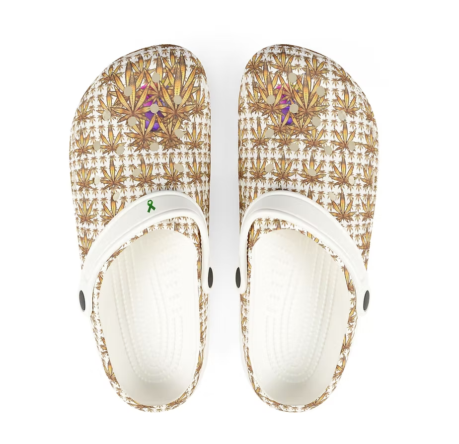 A pair of clogs showcases a vibrant and symmetrical pattern in shades of brown, gold, and white, resembling stylized leaves or abstract shapes. Small purple accents add depth to the design. The clogs feature white straps with black buttons, and a green awareness ribbon is displayed on the left strap. The interior is textured for comfort, blending artistic flair with practical wearability. The overall look is bold and expressive, suggesting a fusion of fashion and advocacy.