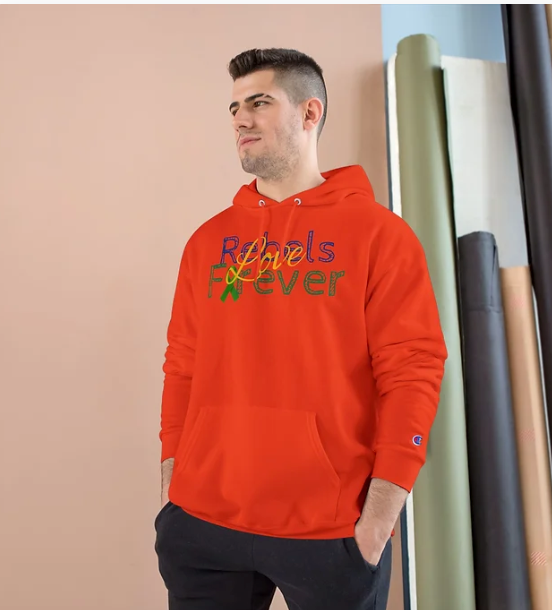 A person stands indoors wearing a bright red hooded sweatshirt with a colorful graphic on the front. The design features the phrase "Rebels Forever"—"Rebels" rendered in a multicolored cursive font and "Forever" in bold green lettering. The background includes a peach-colored wall and several rolled-up fabrics in white, green, and black leaning against a blue wall. The hoodie’s expressive typography and vibrant palette make it a standout piece, blending bold fashion with a spirited message.