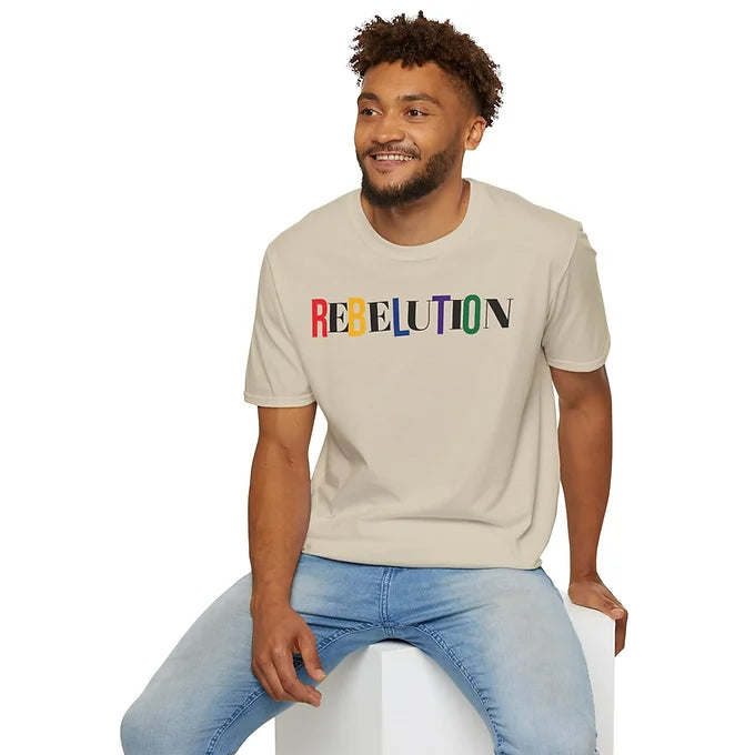 A person sits on a white block wearing a light beige T-shirt with the word "REBELUTION" printed across the chest. Each letter in the word is styled differently, using a mix of colors and fonts that create a visually dynamic and expressive design. Paired with light blue jeans, the overall look is casual yet bold. The shirt’s typography suggests themes of individuality, diversity, and rebellion, making it a standout piece for statement fashion.