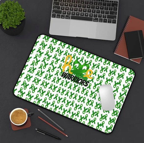 A workspace setup features a laptop, mouse, coffee cup, two pencils, a pen, a small potted plant, and two notebooks. At the center is a mouse pad decorated with a repeating pattern of green awareness ribbons. In the middle, the phrase “hope WARRIORS” appears—“hope” in orange cursive, “WARRIORS” in bold black uppercase, with a green ribbon integrated between the words. The design blends motivation with advocacy, creating a purposeful and empowering workspace vibe.