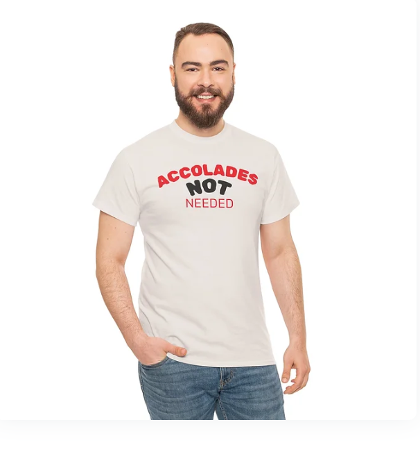 A person stands smiling in a white t-shirt with bold red and black text that reads “ACCOLADES NOT NEEDED.” The design emphasizes humility and self-sufficiency, suggesting the wearer values purpose over praise. The clean layout and confident stance reinforce a message of quiet strength and inner validation.