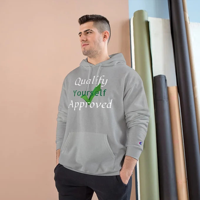 A person wears a light gray hoodie with the motivational phrase “Qualify Yourself Approved” printed on the front. The words “Qualify” and “Approved” are in white, while “Yourself” is in green, matching a green checkmark symbol integrated into the design. The background features a soft pink wall and rolled-up blue and white backdrops, suggesting a studio setting. The hoodie’s message promotes self-validation and personal empowerment.