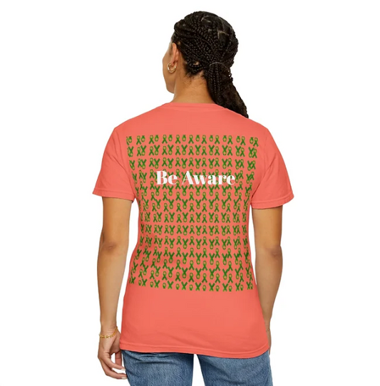 A person is seen from behind wearing a coral-colored T-shirt adorned with rows of green awareness ribbons across the back. In the center of the design, the phrase “Be Aware” is printed in bold white text, standing out against the ribbon pattern. The shirt’s layout is visually striking and symbolic, promoting awareness for causes associated with the green ribbon, such as mental health or organ donation.