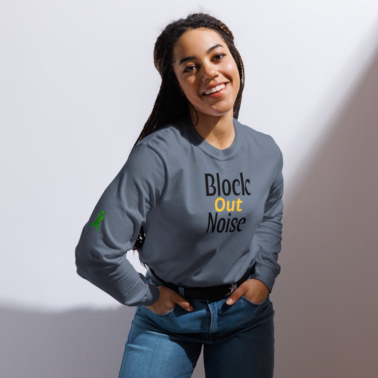 A person smiles while wearing a gray long-sleeve shirt with the phrase “Block Out Noise” printed on the front. The words “Block” and “Noise” are in black, while “Out” is highlighted in yellow. A small green awareness ribbon appears on the left sleeve. The individual stands against a light background with hands resting on their hips, radiating calm and confidence. The shirt’s message encourages focus and resilience, with the ribbon symbol suggesting mental health advocacy.