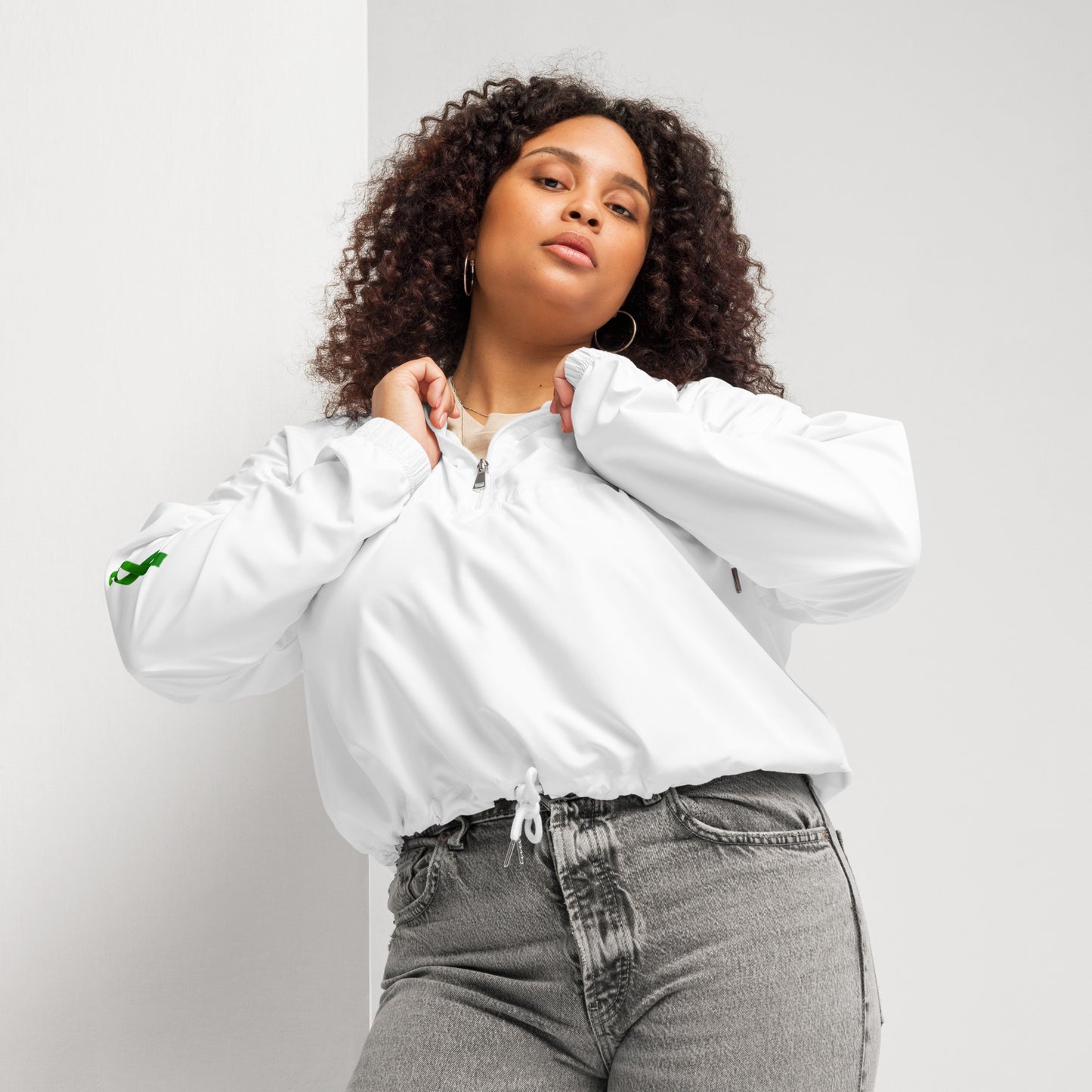A person poses confidently in a white zip-up pullover with a white zipper and drawstring at the waist, paired with high-waisted gray jeans. The pullover features a small green design on the left sleeve. The individual has curly hair and holds the collar with both hands, standing against a plain background. The outfit blends casual comfort with modern style, subtly hinting at advocacy through the green sleeve detail.