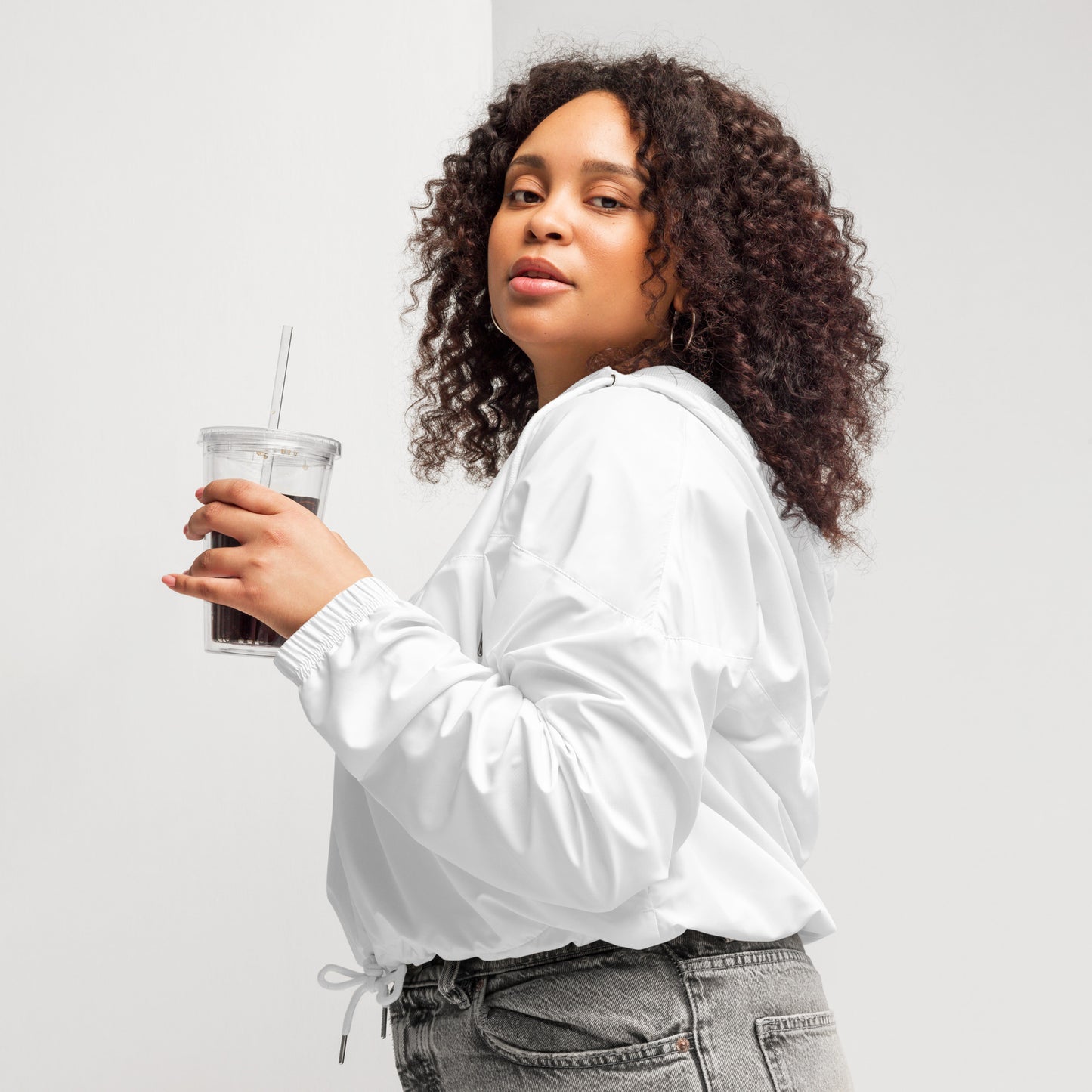 A person poses confidently in a white zip-up pullover with a white zipper and drawstring at the waist, paired with high-waisted gray jeans. The pullover features a small green design on the left sleeve. The individual has curly hair and holds the collar with both hands, standing against a plain background. The outfit blends casual comfort with modern style, subtly hinting at advocacy through the green sleeve detail.