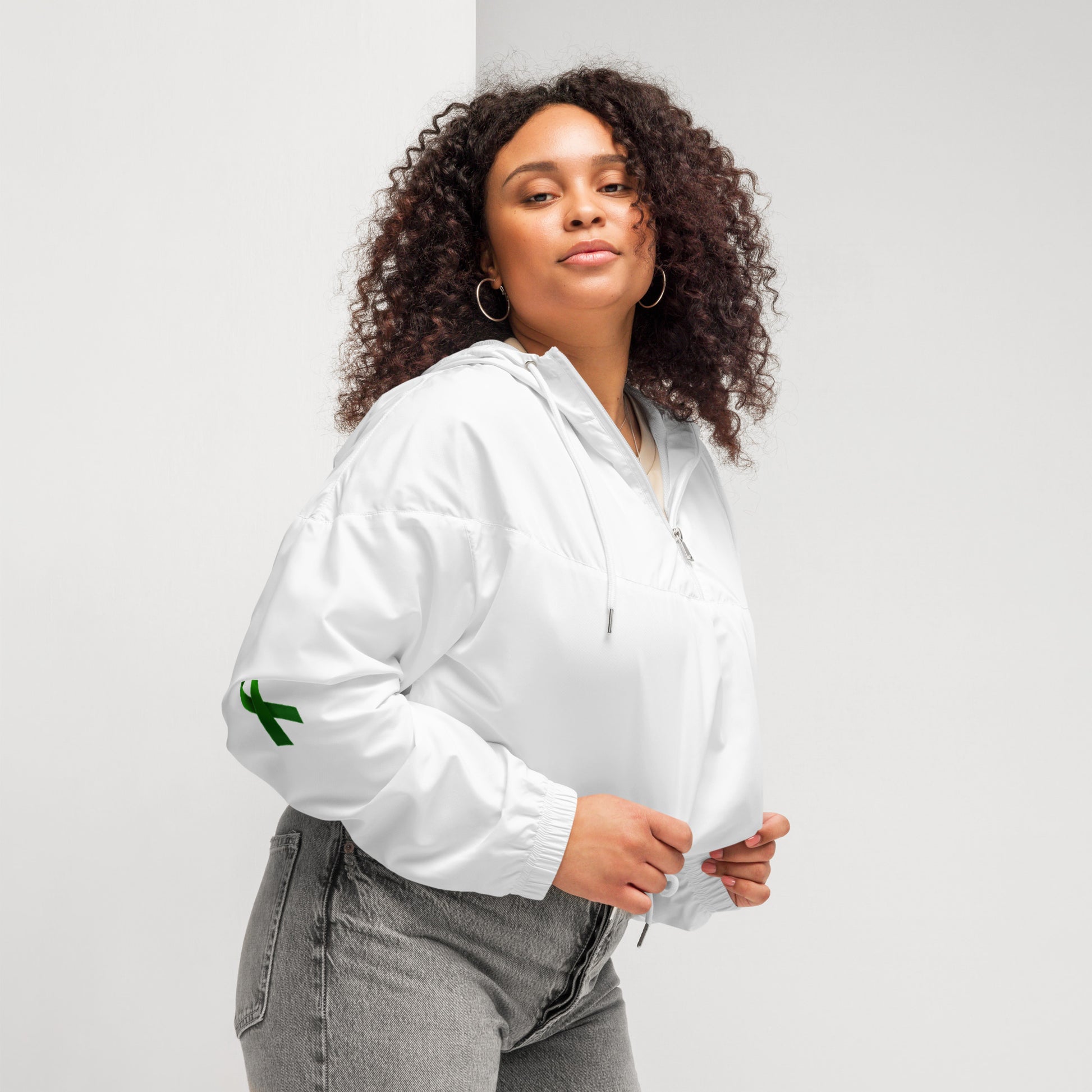 A person poses confidently in a white zip-up pullover with a white zipper and drawstring at the waist, paired with high-waisted gray jeans. The pullover features a small green design on the left sleeve. The individual has curly hair and holds the collar with both hands, standing against a plain background. The outfit blends casual comfort with modern style, subtly hinting at advocacy through the green sleeve detail.