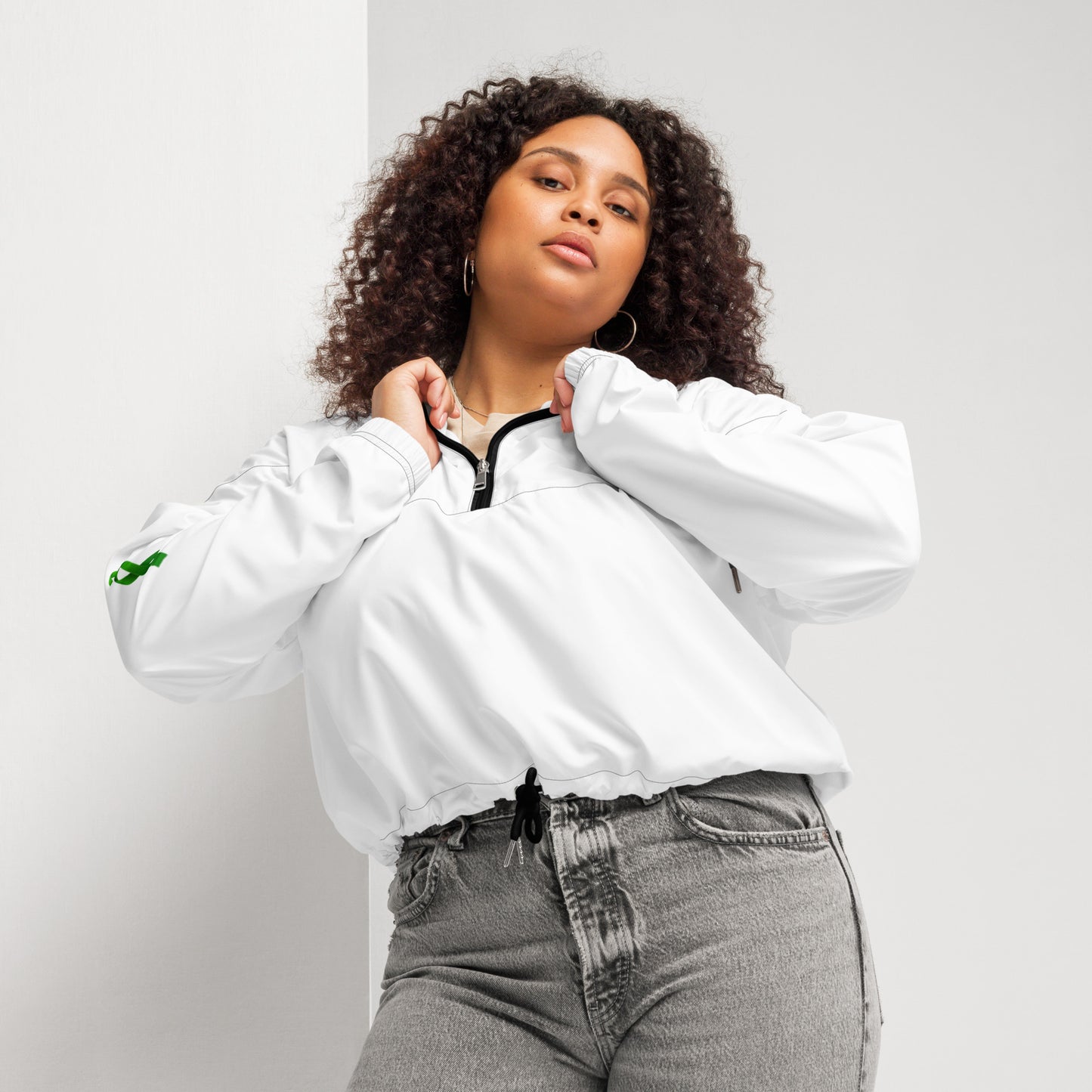 A person poses confidently in a white zip-up pullover with a black zipper and drawstring at the waist, paired with high-waisted gray jeans. The pullover features a small green design on the left sleeve. The individual has curly hair and holds the collar with both hands, standing against a plain background. The outfit blends casual comfort with modern style, subtly hinting at advocacy through the green sleeve detail.