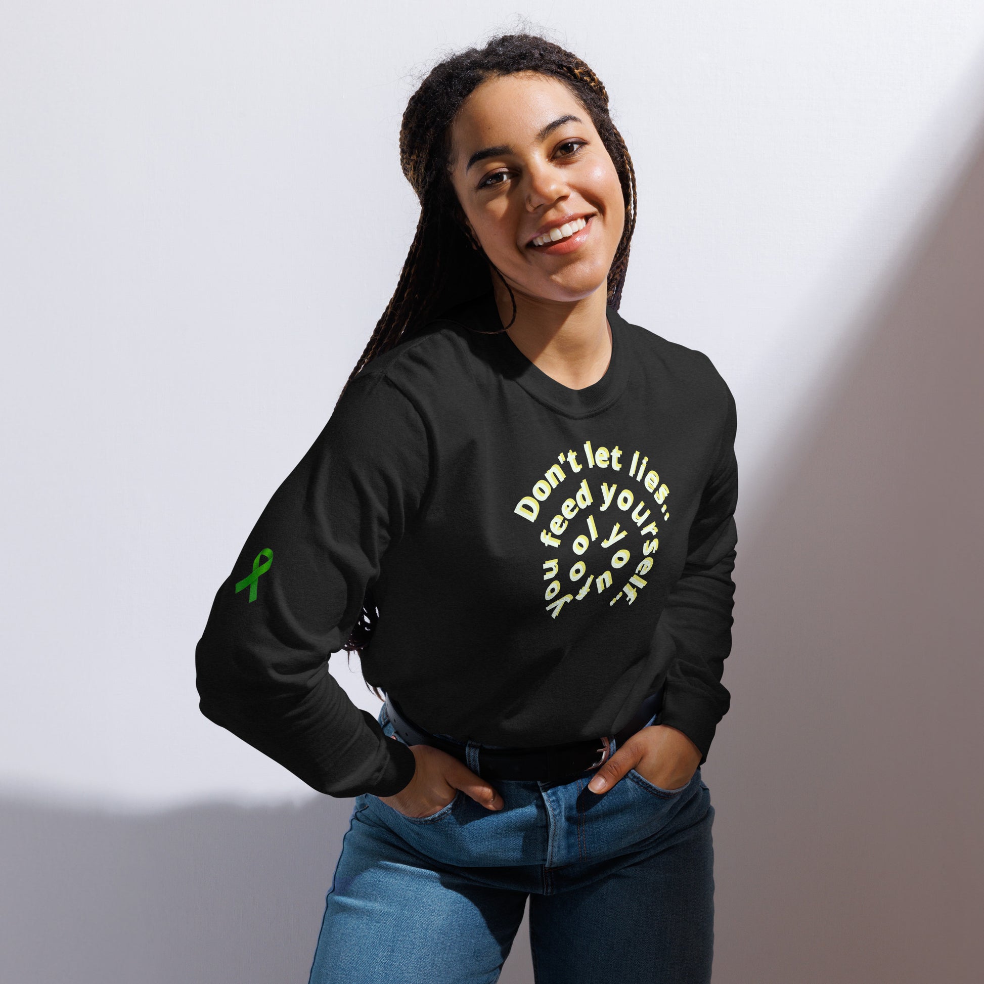 A person stands against a plain background wearing a black long-sleeve shirt with a circular front design that reads “Don’t let illness feed your self-doubt.” A green awareness ribbon is printed on the left sleeve. The shirt’s message promotes mental health encouragement and resilience, blending advocacy with a powerful reminder of inner strength.