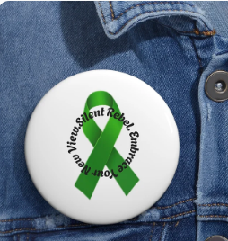 A round white button is pinned to blue denim fabric, featuring a green awareness ribbon at its center. Encircling the ribbon is the phrase “Silent Rebel. Embrace Your New View” in black text. The design blends simplicity with symbolism, promoting mental health awareness and encouraging personal transformation through quiet advocacy.
