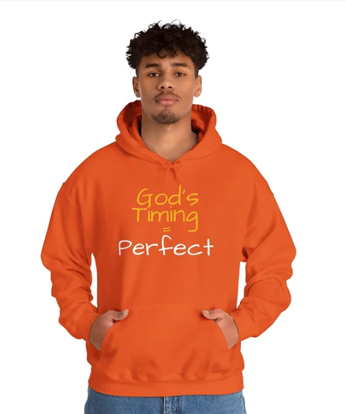 A person wears an orange hoodie with the bold message “God’s Timing = Perfect” printed on the front. The design uses clean typography and vibrant color to convey a faith-based affirmation, emphasizing trust in divine timing. The hoodie blends spiritual encouragement with everyday comfort, making it a wearable reminder of patience and belief.