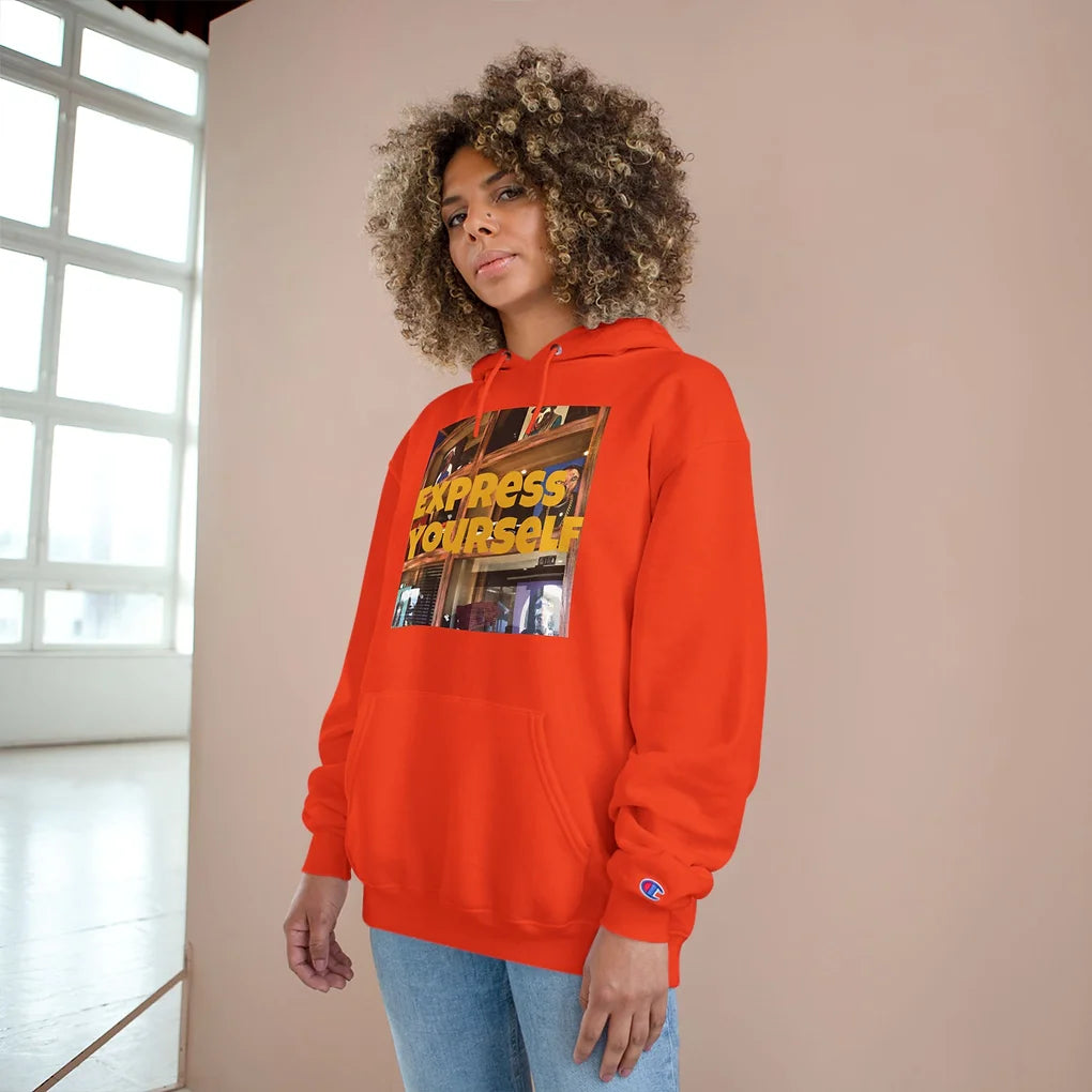 A person stands indoors wearing a bright red hoodie with the bold phrase “EXPRESS YOURSELF” in yellow capital letters across the front. The text overlays a collage of urban and artistic visuals, blending street style with creative energy. A Champion logo appears on the left sleeve. The individual stands against a beige wall with a large window in the background, letting in natural light. The hoodie celebrates individuality, self-expression, and unapologetic creativity.