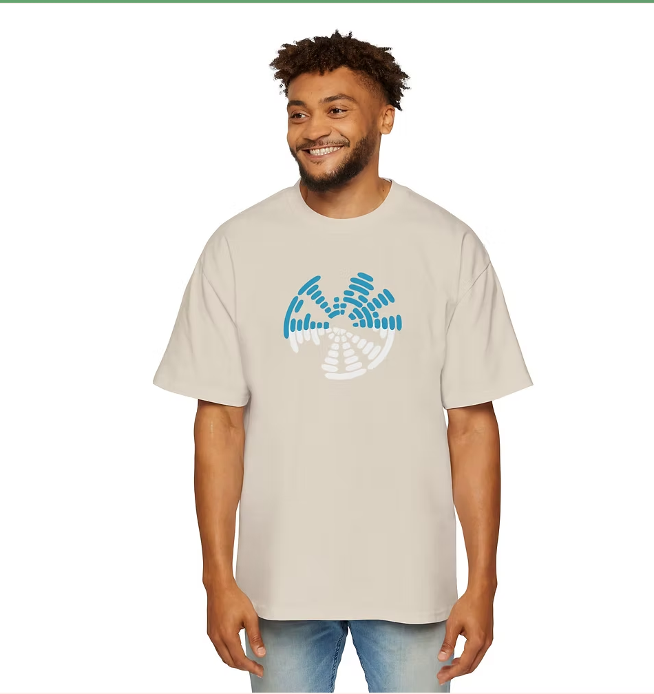 A person wears a beige oversized t-shirt featuring a circular graphic on the front composed of segmented blue and white shapes resembling sound waves or stylized radar signals. Paired with light blue jeans, the look is casual and expressive. The person smiles warmly, standing indoors against a backdrop of rolled-up colorful materials. The design evokes themes of resonance, frequency, and quiet impact—perfect for a campaign centered on subtle strength and emotional clarity.