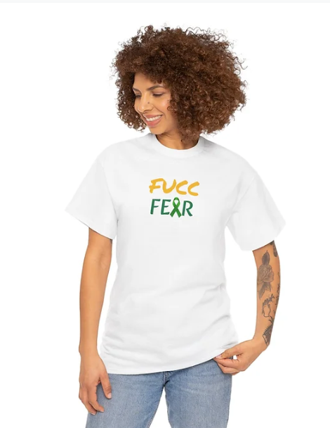 A person wears an white shirt with the bold phrase “FUCC FEAR” printed across the chest. The word “FUCC” is in yellow, green “FEAR” is in white, with the letter “A” replaced by a green awareness ribbon. The shirt is designed to  delivers a defiant message of courage and advocacy, blending bold typography with symbolic support for mental health or related causes.