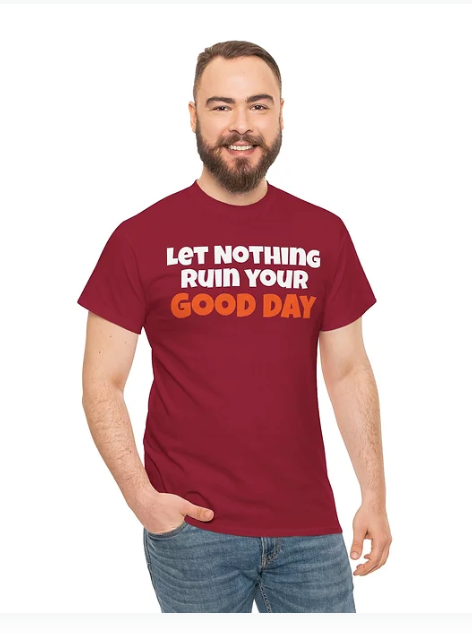 A person stands smiling in a maroon t-shirt with the motivational phrase “LET NOTHING RUIN YOUR GOOD DAY” printed on the front. The words “GOOD DAY” are highlighted in orange, while the rest of the text appears in white. The design radiates positivity and resilience, offering a wearable reminder to protect joy and stay grounded in optimism.