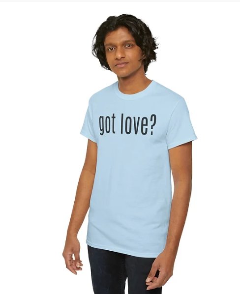 A person stands wearing a light blue t-shirt with the phrase “got love?” printed in black across the chest. The design plays on familiar advertising language to pose a reflective, heart-centered question. Paired with casual blue jeans and long, curly hair, the look blends simplicity with emotional resonance—inviting connection, compassion, and quiet advocacy.