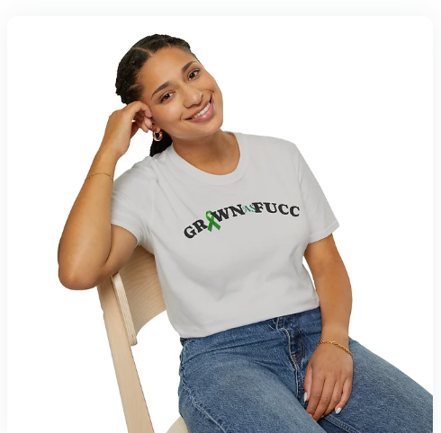 A person sits smiling on a wooden chair, wearing a white t-shirt with the bold phrase “GROWNFUCC” printed across the chest. The asterisk is stylized as a green awareness ribbon, blending defiance with advocacy. Paired with blue jeans, hoop earrings, and a nose ring, the look radiates unapologetic maturity, resilience, and cause-driven identity. The plain white background keeps the focus on the message and the person’s confident presence.*