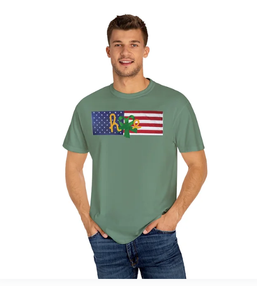 A man wears a green t-shirt featuring a stylized American flag design with the word “hope” creatively integrated. The “o” is shaped like a sunflower, and the “p” incorporates a green awareness ribbon. The design blends patriotic symbolism with emotional advocacy, offering a message of resilience, mental health awareness, and national unity.