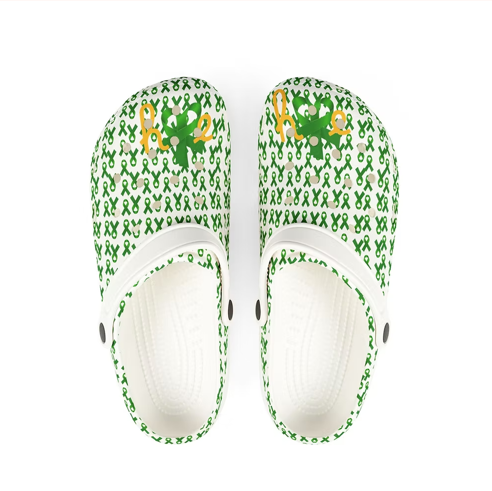 A pair of white clogs are covered in a pattern of green awareness ribbons. Each clog features a cheerful green character with a mustache and hat, resembling a leprechaun, alongside the word “HOPE” in bold yellow and green letters. The interior is black with a textured footbed. The design blends playful charm with advocacy, offering a whimsical yet powerful message of support and visibility.