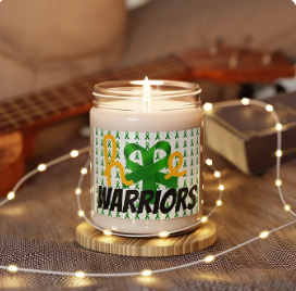 A lit candle in a glass jar sits on a wooden coaster, surrounded by string lights, a book, and a guitar in the background—creating a warm, cozy ambiance. The candle’s label features the word “HOPE,” with the “O” replaced by a green ribbon shaped like a heart, and the word “WARRIORS” boldly printed below. The background of the label is decorated with small green awareness ribbons, blending comfort with advocacy and emotional strength.