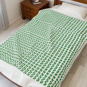 A neatly made bed features a white blanket patterned with repeating green ampersands (“&”) arranged in diagonal rows. The design is bold and uniform, standing out against the room’s minimalist decor, which includes light wood flooring, a wooden headboard, and a matching nightstand with a book and small object on top. The blanket’s visual rhythm suggests connection, continuation, and quiet advocacy.