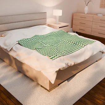A neatly made bed features a white blanket patterned with repeating green ampersands (“&”) arranged in diagonal rows. The design is bold and uniform, standing out against the room’s minimalist decor, which includes light wood flooring, a wooden headboard, and a matching nightstand with a book and small object on top. The blanket’s visual rhythm suggests connection, continuation, and quiet advocacy.