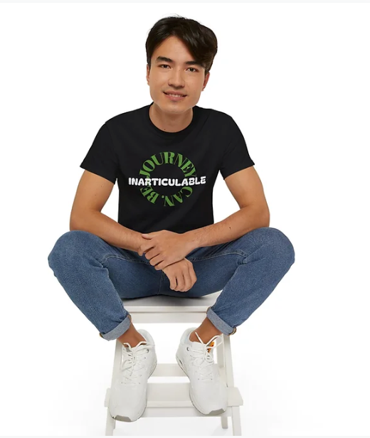 A person sits cross-legged on a white stool in front of a plain white background, wearing a black t-shirt with a striking design. The shirt features the phrase "JOURNEY CAN BE INARTICULABLE"—with "JOURNEY CAN BE" arranged in a circular green font and "INARTICULABLE" boldly printed in white across the center. The individual pairs the shirt with blue jeans and white sneakers, creating a casual yet thought-provoking look that emphasizes introspection and the complexity of personal experiences.