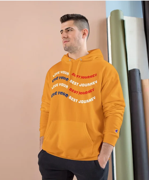 A person wears a bright orange Champion hoodie featuring a wave-like graphic on the front. The design repeats the phrase "LIVE YOUR BEST JOURNEY" four times, with "LIVE YOUR" alternating in white and dark blue, and "BEST JOURNEY" in white and red. The layered typography creates a rhythmic, motivational visual. The background includes a peach-colored wall and rolled-up backdrops in green, black, and white, suggesting a studio setting. The hoodie’s vibrant color and uplifting message make it a bold choice for