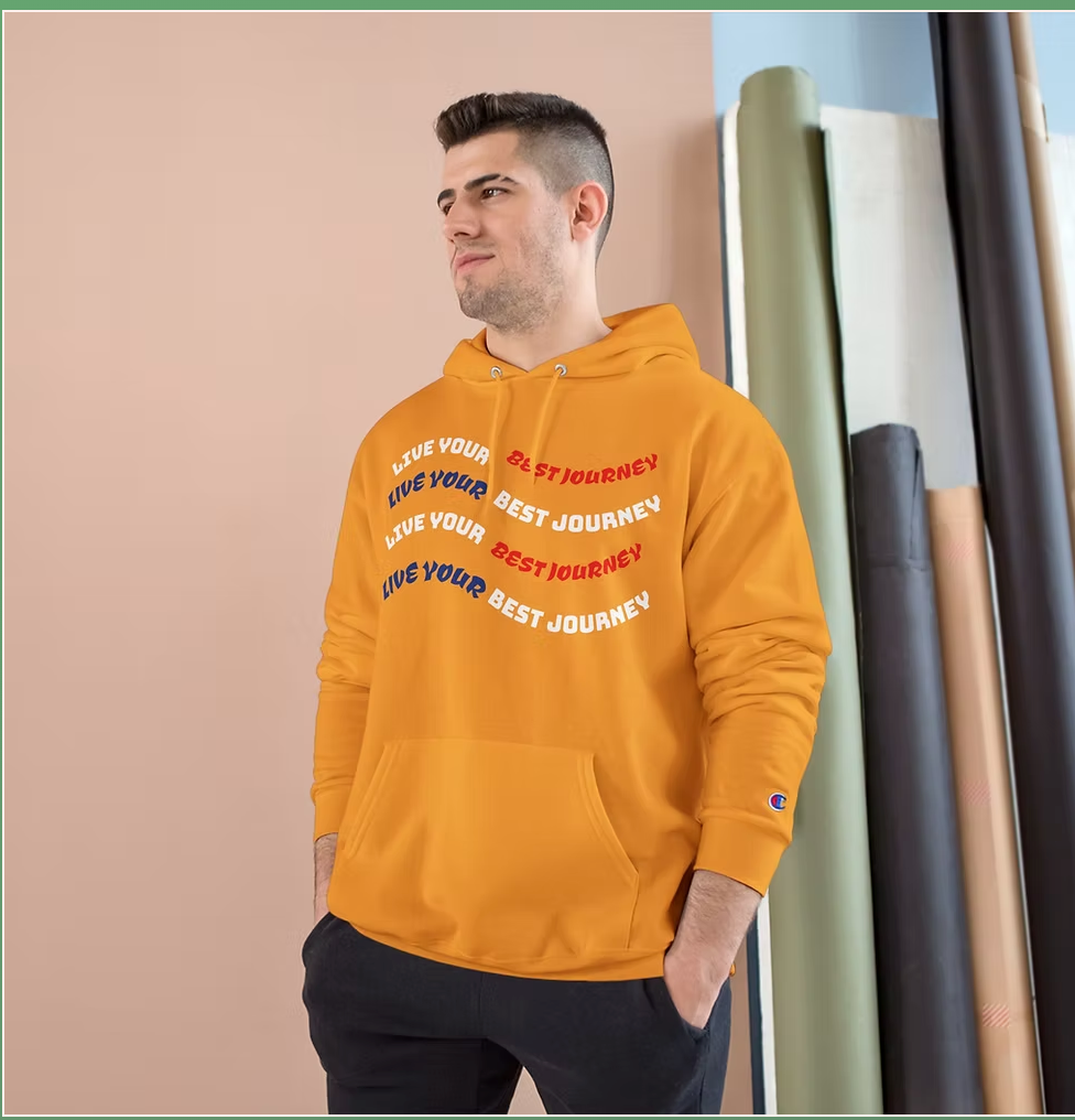 A person stands in front of a peach-colored wall wearing a vivid orange Champion hoodie. The hoodie features the motivational phrase "LIVE YOUR BEST JOURNEY" repeated four times in a wave-like pattern across the chest, alternating in white, blue, and red text. Behind the person are several rolled-up fabric backdrops in various colors, suggesting a creative studio environment. The overall vibe is energetic and uplifting, with a strong emphasis on positivity and bold fashion.
