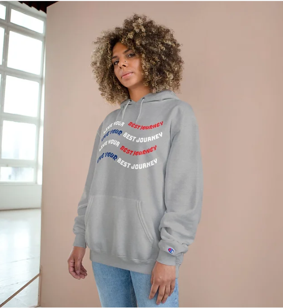 A person wears a grey Champion hoodie featuring a motivational message printed in a curved, repeating pattern across the chest. The phrase "LOVE YOUR BEST JOURNEY" appears multiple times, with "LOVE YOUR" in white and blue, and "BEST JOURNEY" alternating in white and red. The Champion logo is visible on the left sleeve cuff. The background includes a beige wall and a window letting in natural light, creating a soft indoor ambiance. The design blends inspiration with casual style, ideal for everyday wear wit