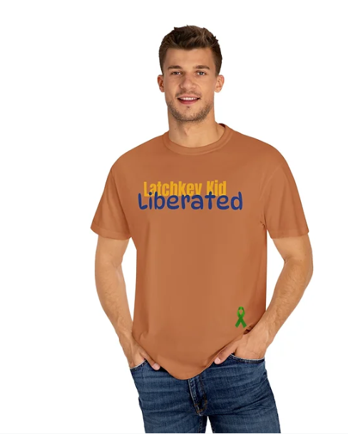 A man stands casually with his hands in his pockets, wearing a brown T-shirt that reads "Latchkey Kid Liberated." The phrase is split by color: "Latchkey Kid" in yellow and "Liberated" in blue. A small green ribbon graphic appears near the lower right side of the shirt. He pairs the tee with jeans, and the neutral background suggests an indoor setting. The design conveys a message of empowerment and individuality, possibly referencing the experience of growing up independently.