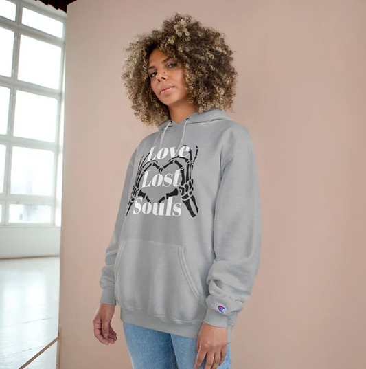 A person wears a gray Champion hoodie featuring a bold graphic on the front: two skeletal hands forming a heart shape, with the phrase "Love Lost Souls" printed in large white letters across the design. The background includes a beige wall and a window letting in soft natural light, creating a calm indoor setting. The hoodie’s artwork evokes a mix of gothic style and emotional depth, blending themes of love and mortality.