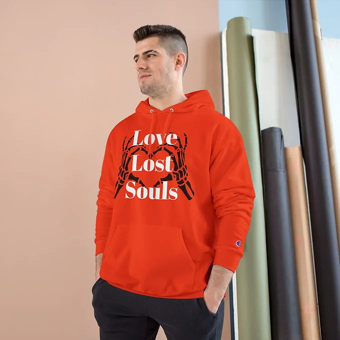 A person wears a orange Champion hoodie featuring a bold graphic on the front: two skeletal hands forming a heart shape, with the phrase "Love Lost Souls" printed in large white letters across the design. The background includes a beige wall and a window letting in soft natural light, creating a calm indoor setting. The hoodie’s artwork evokes a mix of gothic style and emotional depth, blending themes of love and mortality.