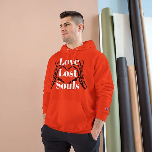 A person wears a orange Champion hoodie featuring a bold graphic on the front: two skeletal hands forming a heart shape, with the phrase "Love Lost Souls" printed in large white letters across the design. The background includes a beige wall and a window letting in soft natural light, creating a calm indoor setting. The hoodie’s artwork evokes a mix of gothic style and emotional depth, blending themes of love and mortality.