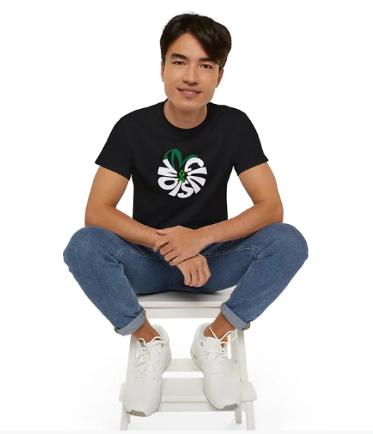 A person sits cross-legged on a white stool against a plain white background, wearing a black T-shirt with a bold graphic design. The front of the shirt features the word "NOISE" arranged in a circular pattern using stylized white letters, intertwined with a green element resembling a leaf or ribbon. The individual pairs the shirt with rolled-cuff blue jeans and white sneakers, presenting a relaxed and modern look. The design stands out for its artistic flair and subtle symbolism, possibly hinting at awaren