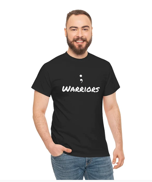 A person wears a black T-shirt with the phrase "; WARRIORS" printed in bold white stylized font. The semicolon symbol is prominently placed above the word "WARRIORS," creating a powerful visual statement. The design is simple yet impactful, often associated with mental health awareness and the idea of perseverance—symbolizing strength, survival, and the choice to continue. The overall look is minimalistic and meaningful, resonating with themes of resilience and hope.