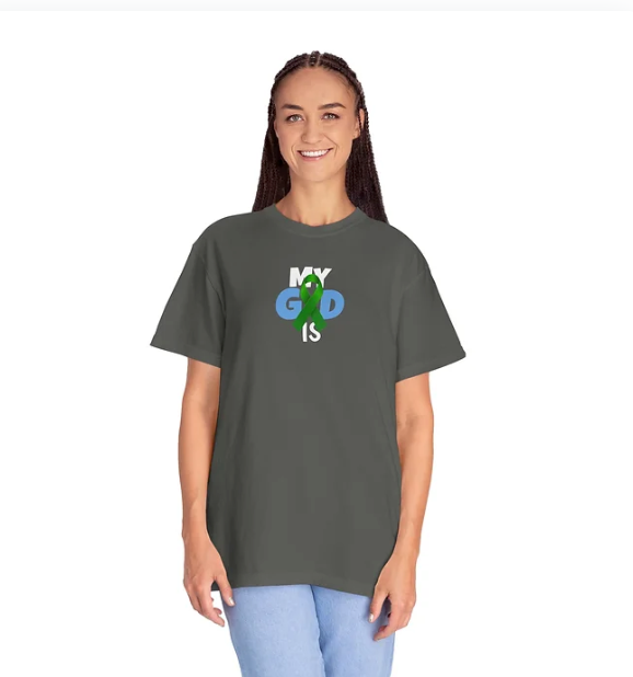 A person wears a dark-colored T-shirt with the phrase "MY GOD IS" printed across the chest in a stylized font. The word "GOD" is creatively illustrated—its "O" replaced by an image of the Earth in green and blue, while the letters "G" and "D" are in blue and the rest of the text is in white. The person is smiling and dressed in light blue pants, standing against a plain background. The design blends spiritual messaging with a global theme, suggesting the universality and omnipresence of faith.