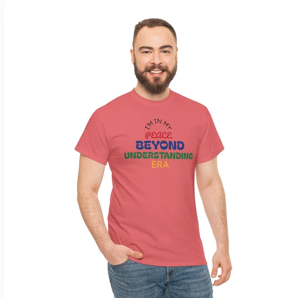 A person stands smiling in a red T-shirt with a multicolored text design that reads: "I'M IN MY peace BEYOND UNDERSTANDING ERA." The phrase is arranged with varied font sizes and colors to emphasize key words like "peace" and "UNDERSTANDING." The individual pairs the shirt with blue jeans and stands against a plain white background. The design conveys a message of personal growth, tranquility, and empowerment, making it both expressive and motivational.