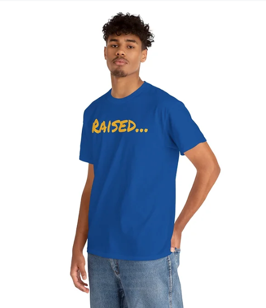 A person stands casually in a royal blue T-shirt with the word "RAISED..." printed across the chest in bold, stylized yellow letters. The phrase ends with ellipses, inviting curiosity and personal interpretation. Paired with casual blue jeans and a relaxed pose—one hand in a pocket—the look is simple yet impactful. The shirt’s design uses contrast and ambiguity to spark conversation and express individuality.