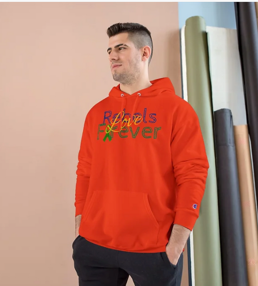 A person stands indoors wearing a bright red hooded sweatshirt with a colorful graphic on the front. The design features the phrase "Rebels Forever"—"Rebels" rendered in a multicolored cursive font and "Forever" in bold green lettering. The background includes a peach-colored wall and several rolled-up fabrics in white, green, and black leaning against a blue wall. The hoodie’s expressive typography and vibrant palette make it a standout piece, blending bold fashion with a spirited message.
