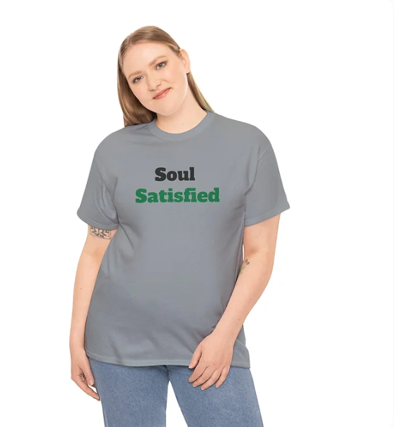 A person stands wearing a gray T-shirt with the phrase "Soul Satisfied" printed across the chest. The word "Soul" appears in black, while "Satisfied" is rendered in green, creating a subtle yet expressive contrast. The individual poses with a slight tilt, one hand resting on the hip and the other relaxed. The shirt’s message evokes themes of inner peace and contentment, making it a meaningful choice for casual, motivational fashion.