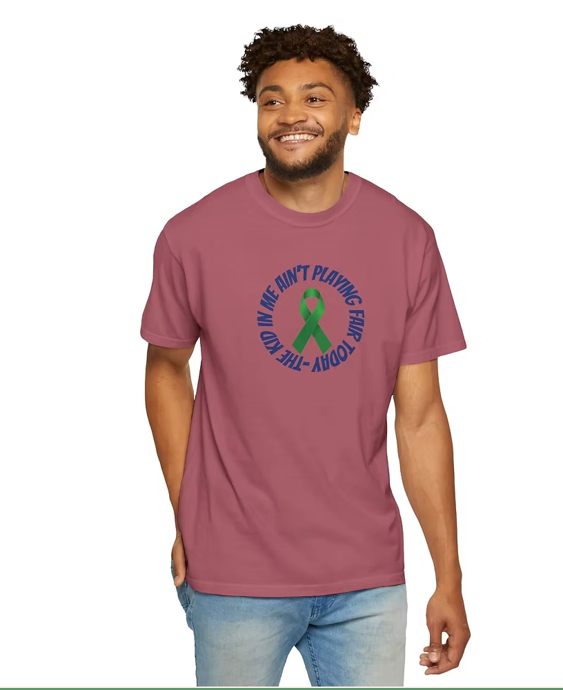 A person wears a maroon T-shirt featuring a bold graphic design. At the center is a green and red awareness ribbon, surrounded by blue text that reads: "THE KID AIN'T PLAYING FAIR TODAY." The shirt’s message conveys a strong, assertive attitude, while the ribbon suggests a connection to advocacy or a cause. The combination of expressive typography and symbolic imagery makes this piece both visually impactful and potentially meaningful.
