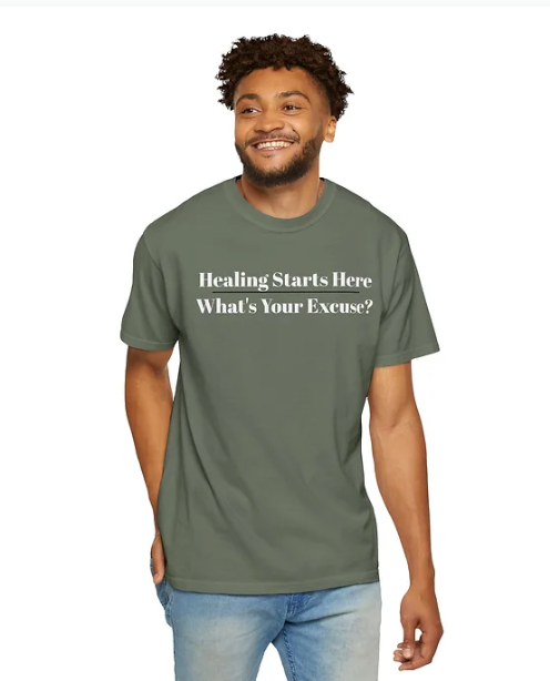 A person stands smiling in a dark green T-shirt with white text printed on the front. The shirt reads: "Healing Starts Here" on the first line, followed by "What's Your Excuse?" beneath it. One hand is tucked into a pocket, and the individual wears a casual, confident expression. The message is motivational and direct, encouraging personal growth and accountability through bold, minimalist typography.