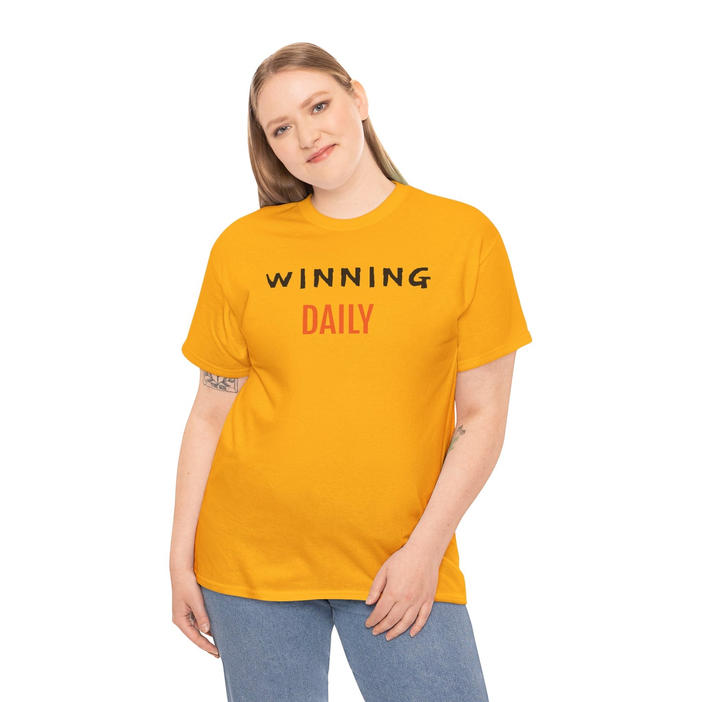 A person wears a bright orange T-shirt with the phrase "WINNING DAILY" printed boldly across the front. "WINNING" is styled in black uppercase letters, while "DAILY" appears in red uppercase letters beneath it. The individual stands with a relaxed posture, revealing tattoos on both forearms. Set against a plain white background, the shirt’s design delivers a confident and motivational message, ideal for lifestyle branding or expressive fashion.